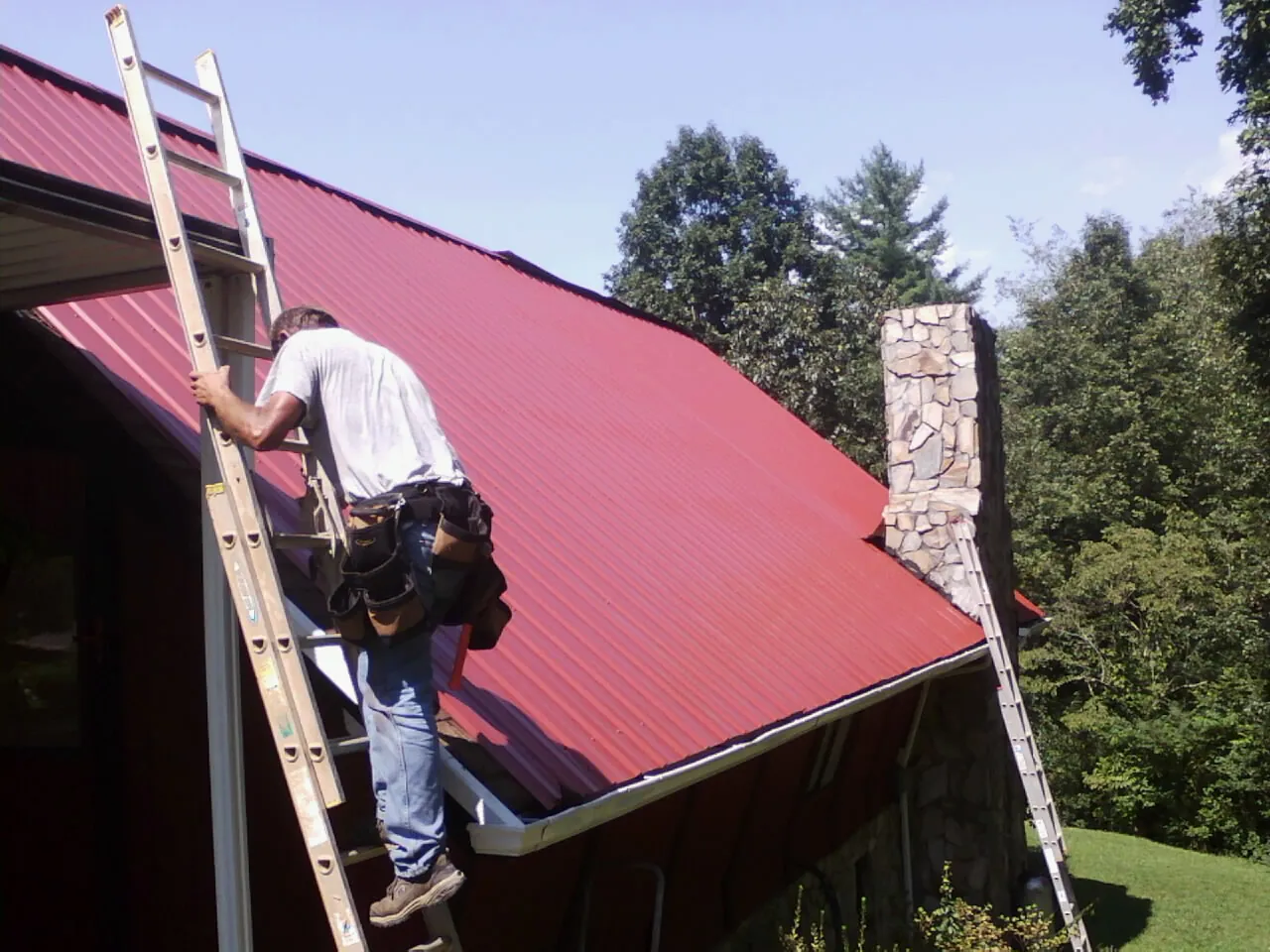 Roofer on a ladder beside a red metal roof and stone chimney — Soffit & Fascia Repair by Roofing Brothers in Lacrosse